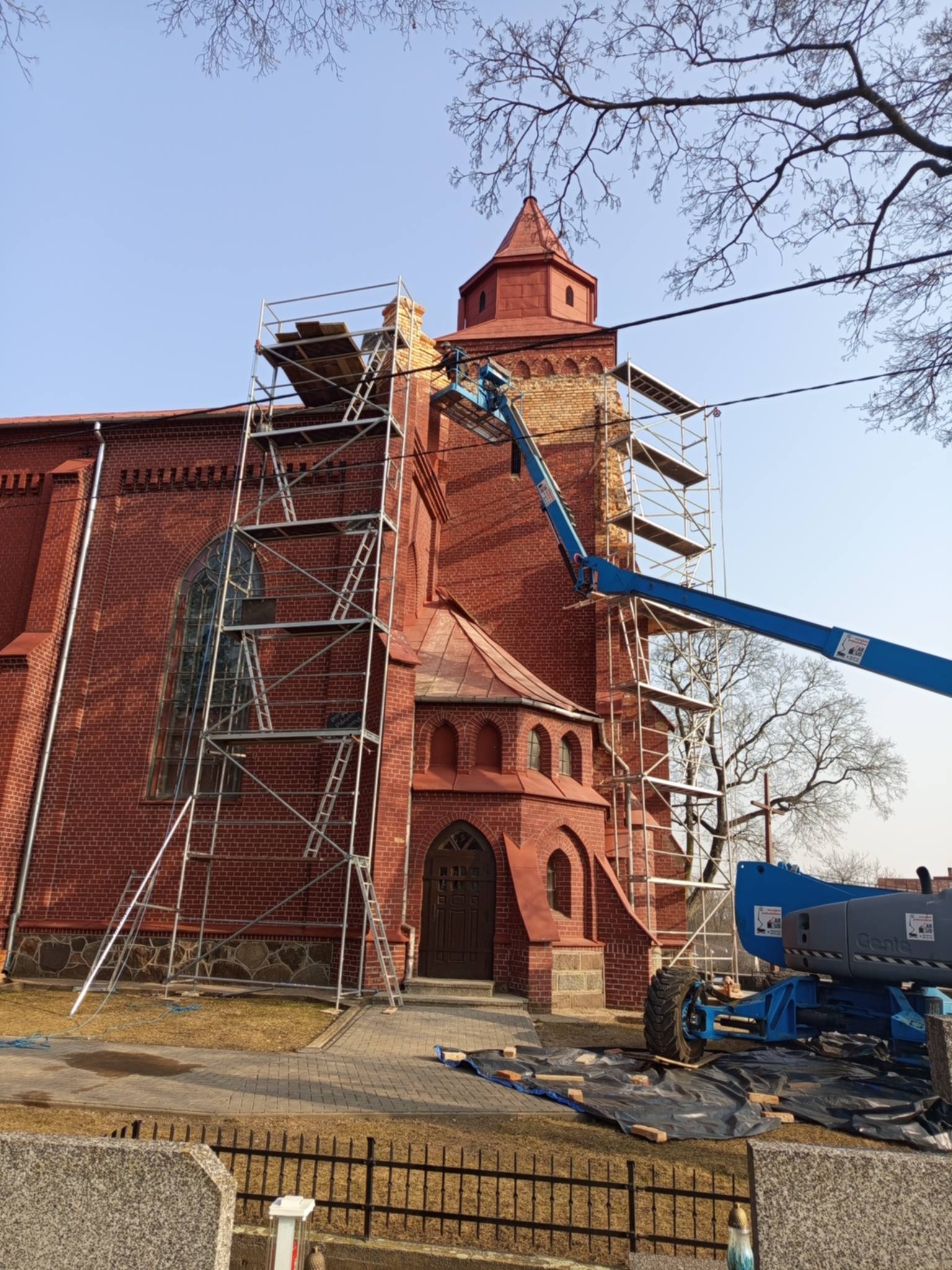 Prace remontowe przy elewacji kościoła w Wyszynach Kościelnych, robotnicy na podnośniku wymieniają cegły.